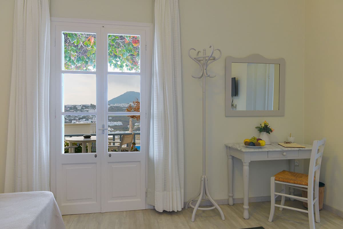 A bright hotel room with white doors opening onto a balcony with a view of the hills