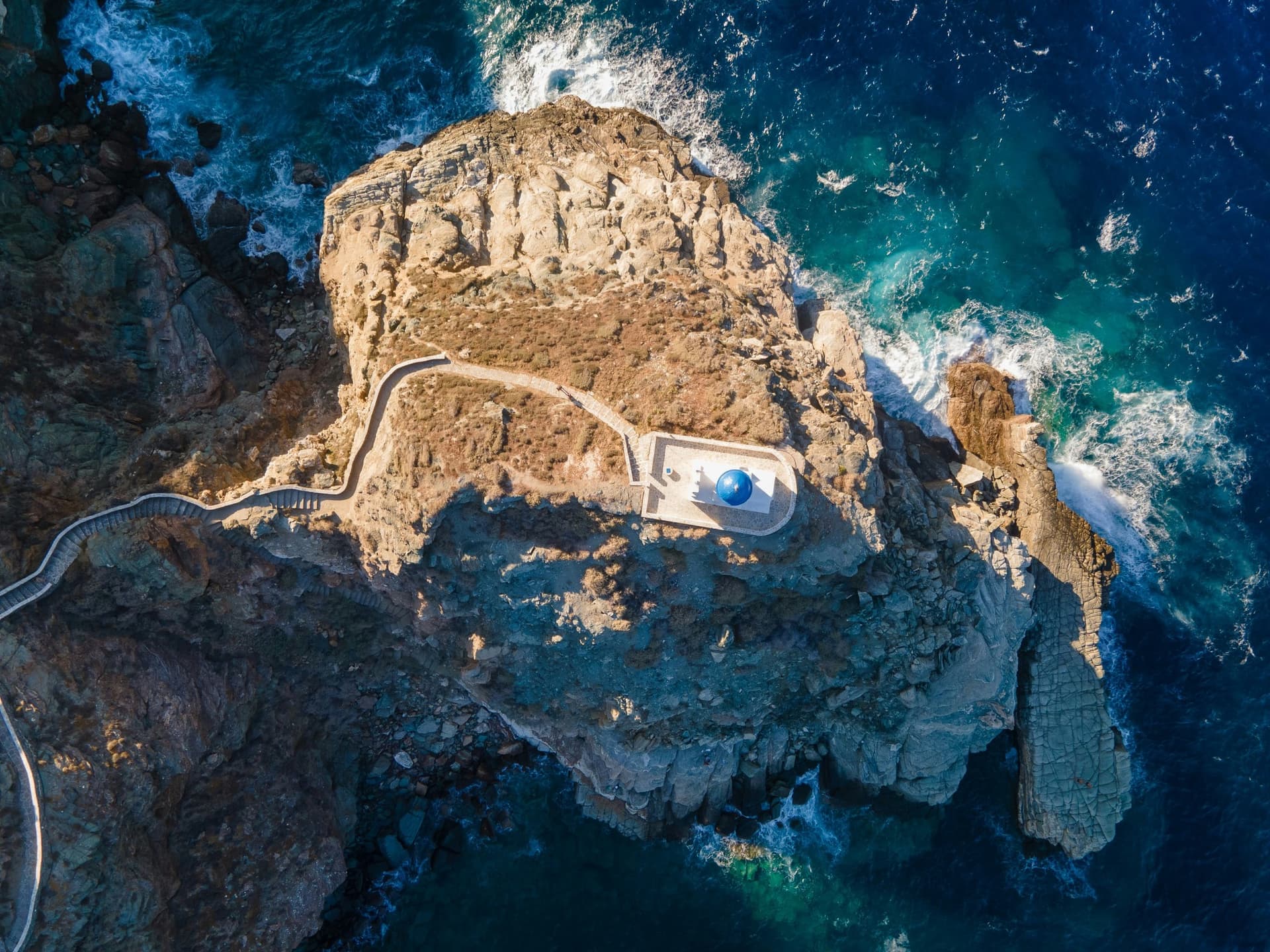 Drone view straight down on the Seven Martyrs chapel on its rock, surrounded by deep teal sea and surf