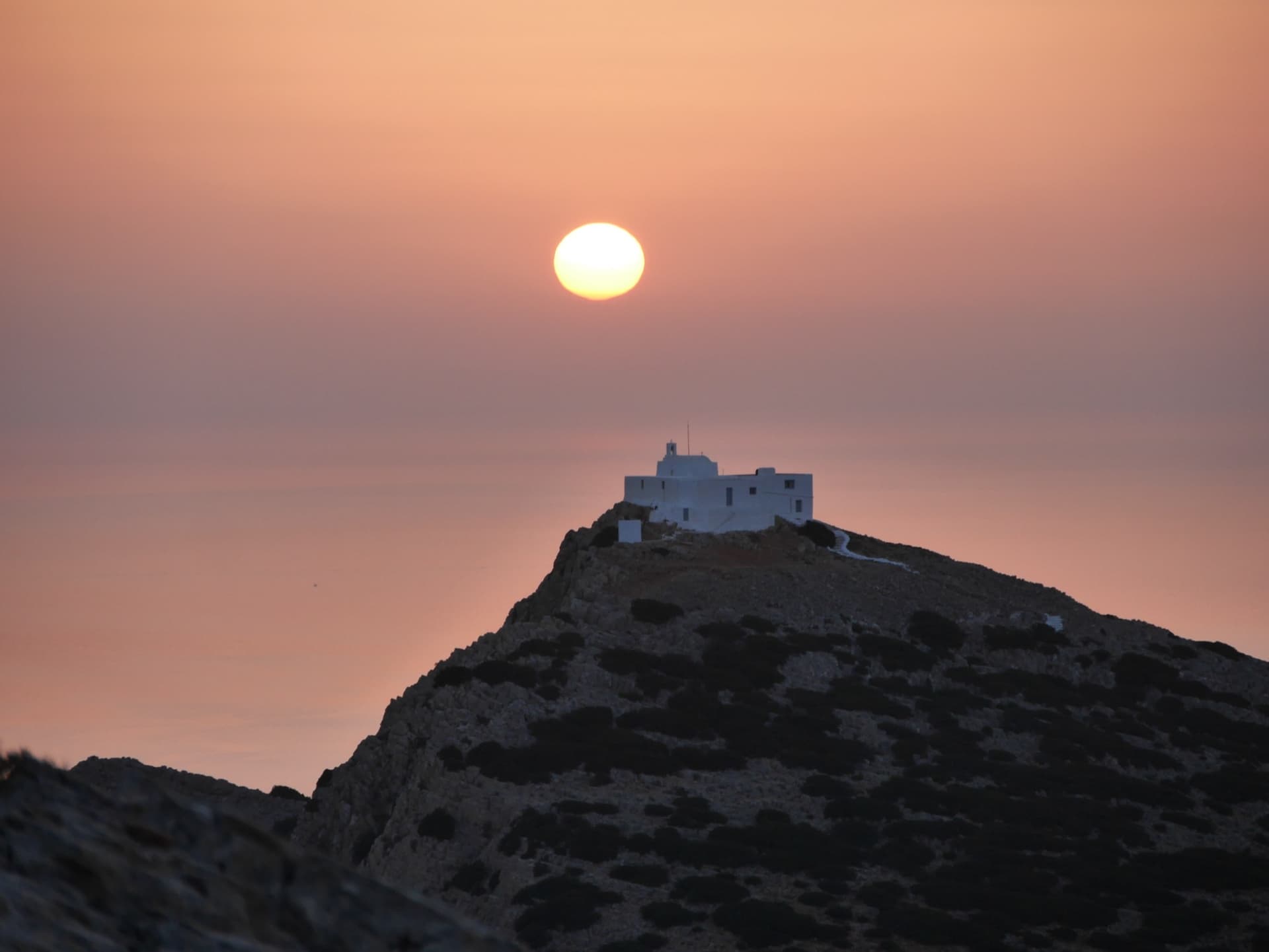 A whitewashed chapel on a hilltop silhouetted against a huge orange setting sun